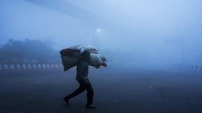 New Delhi: A man carrying sacks of goods walks by amid dense fog on a winter morning, at Mayur Vihar area, in New Delhi, Saturday, Dec. 20, 2025. Visibility plunged across parts of Delhi on Saturday morning, with buildings and flyovers fading into a grey blur amid dense fog, as the capitals AQI edged closer to the severe  category at 384. (PTI Photo/Ravi Choudhary) (PTI12_20_2025_000036B)