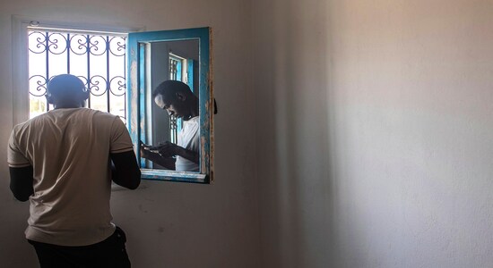 In this Monday, Sept. 23, 2019 photo, Abdullah, 25, a Sudanese migrant who tried crossing the Mediterranean from Libya, is reflected in a window as he uses his phone inside a Tunisian Red Crescent facility in Zarzis, southern Tunisia. The group of 47 in his first crossing from Tripoli over a year earlier had paid a uniformed Libyan and his cronies $127,000 in a mix of dollars, euros and Libyan dinars for the chance to leave their detention center and cross in two boats. They were intercepted in a coast guard boat by the same uniformed Libyan, shaken down for their cell phones and more money, and tossed back into detention. (AP Photo/Mosa'ab Elshamy)